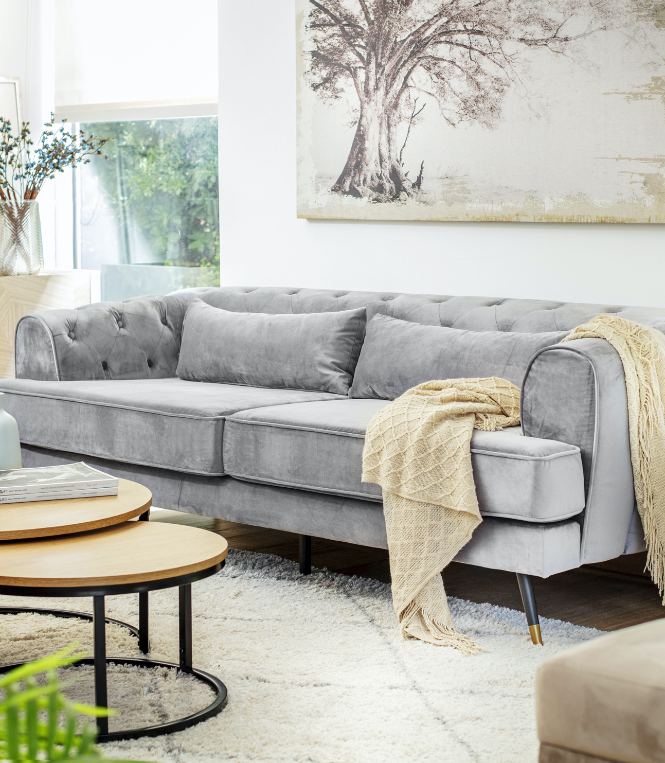 Living de paredes blancas con un gran cuadro en tonos crema, un sofá gris estilo capitoné con una manta color arena encima. Frente al sofá hay 2 mesas de centro redondas que se sobreponen.