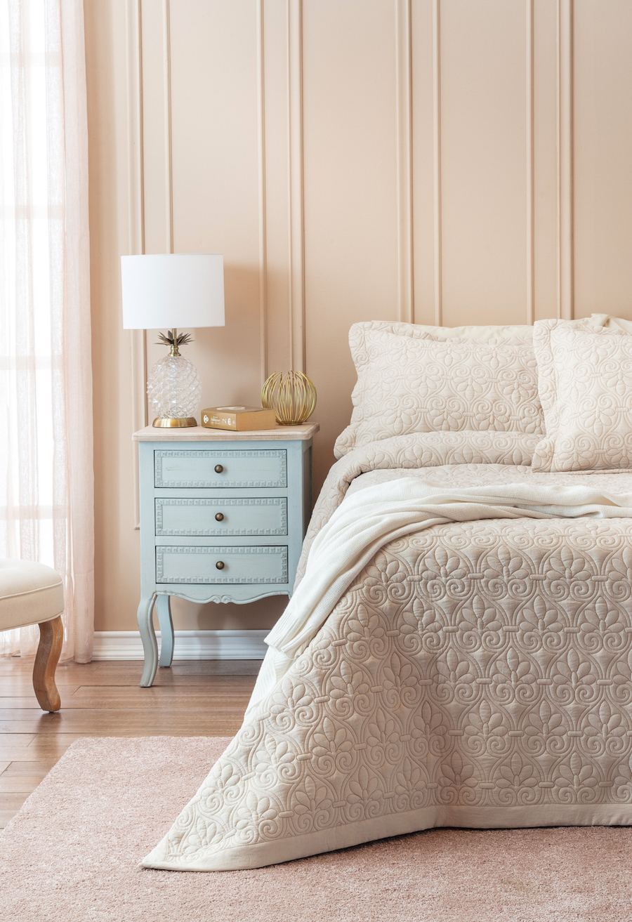 Dormitorio clásico con pared rosa con molduras. Cama con cobertor beige, velador celeste vintage y lámpara de mesa.