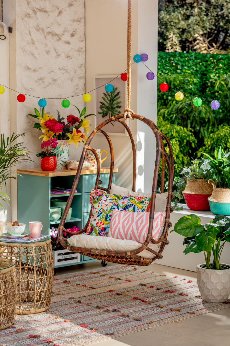 Terraza con silla colgante, con cojines de varios colores. Guirnaldas colgantes de colores, mueble celeste de madera con plantas y adornos. Pequeña mesa de apoyo de bambú, con bandeja y vajilla.