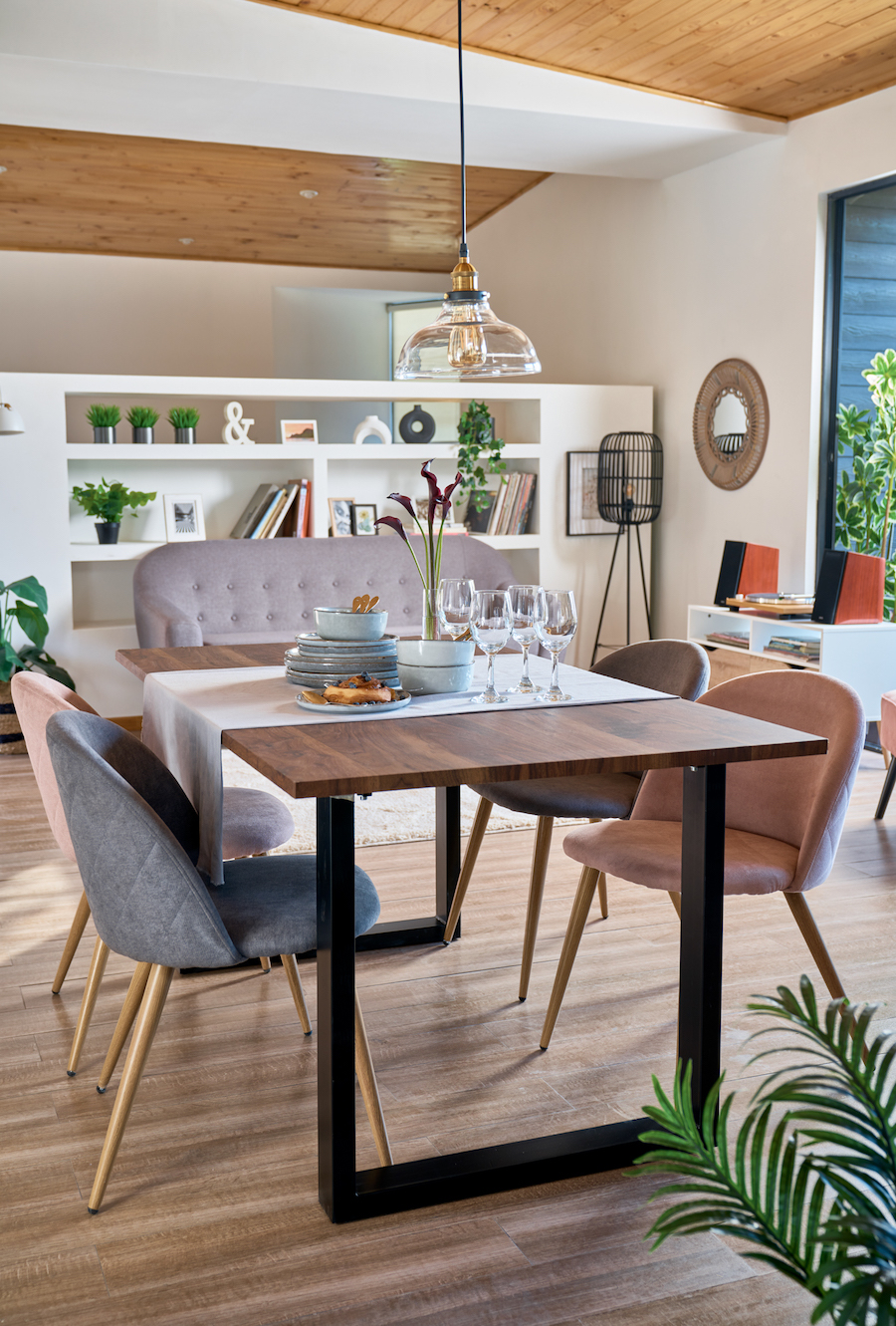 Living comedor moderno con mesa de madera y cuatro sillas en tonos grises, beige y café. Atrás hay un sofá gris y una gran repisa mural con adornos y libros.