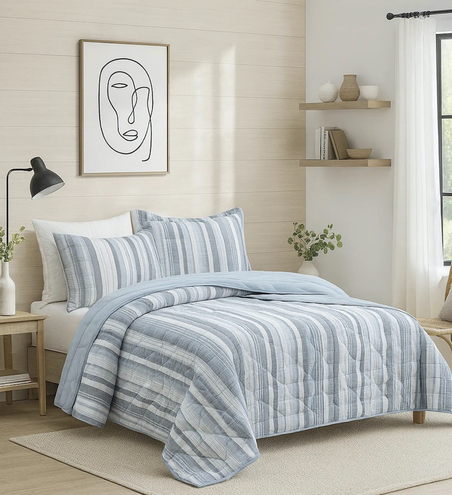 Dormitorio de estilo Nórdico con una cama con un quilt de líneas blancas y azul claro. Sobre la cama hay un cuadro con fondo blanco y líneas blancas, con marco de madera clara. También hay unas repisas con adornos en tonos neutros.