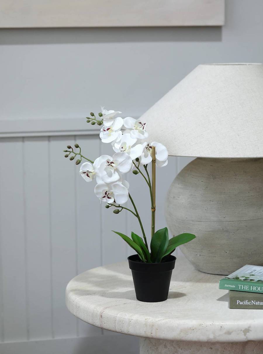 Detalle de una mesa lateral en un living con paredes grises. Sobre la mesa hay una lámpara redonda gris con pantalla blanca, una pila de libros y una orquídea artificial en un macetero negro.