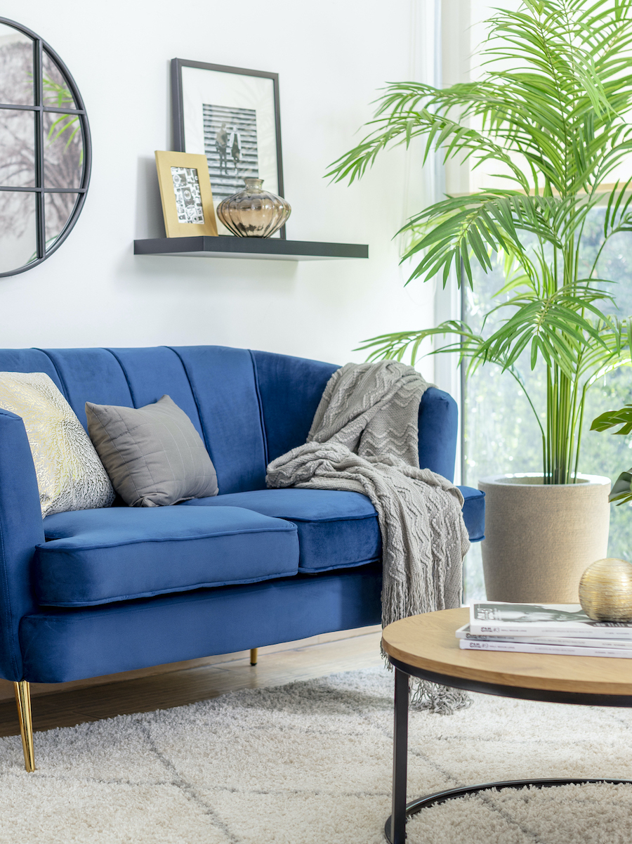 Sala de estar con paredes blancas y piso de madera clara, con un sofá de 2 cuerpos azul con cojines en tonos grises y una alfombra shaggy en los mismos tonos. Junto al sofá hay una gran planta artificial dentro de un macetero beige.