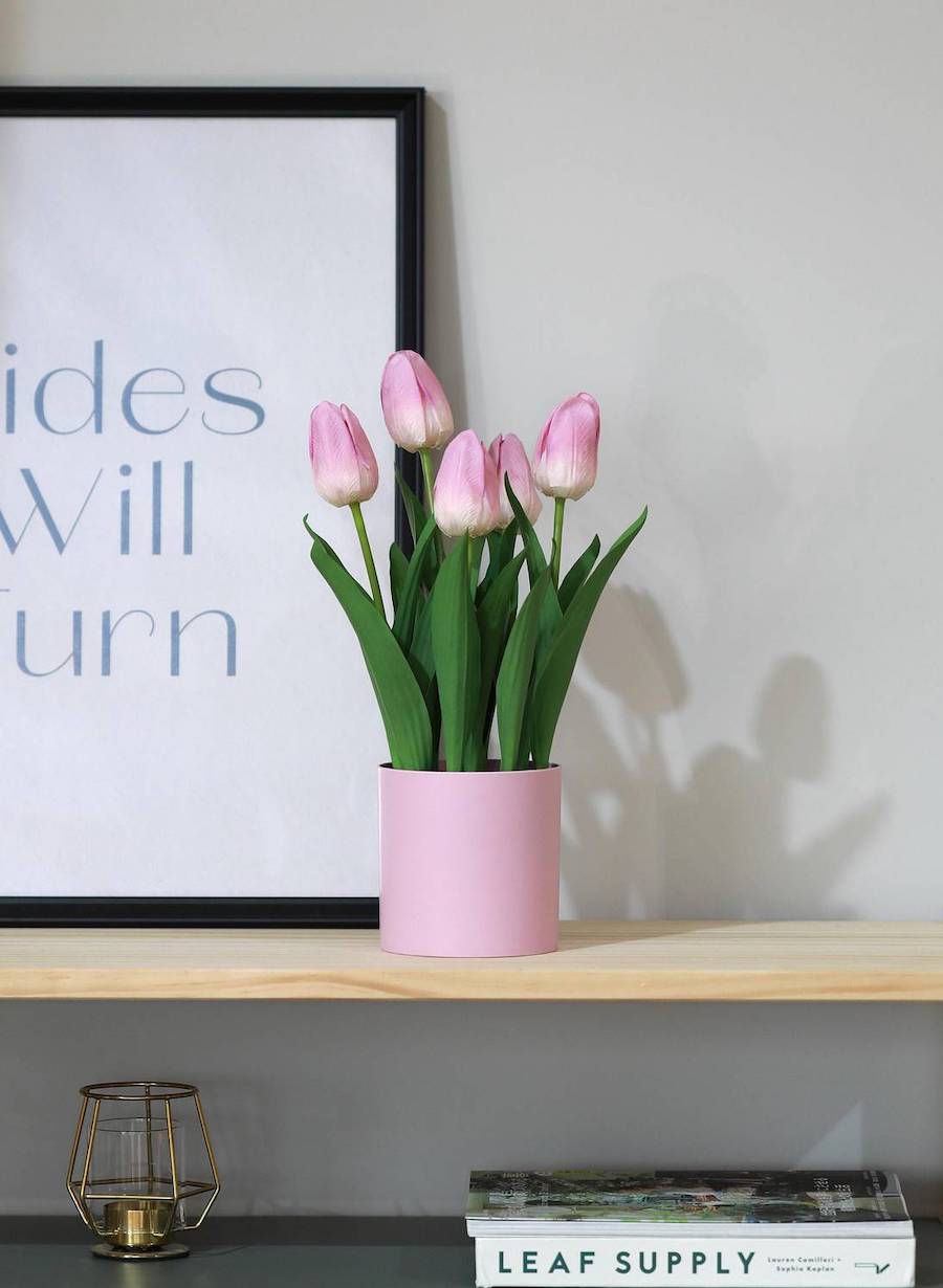 Repisa de madera clara con un cuadro con una frase junto a un macetero rodado con tulipanes artificiales rosados.