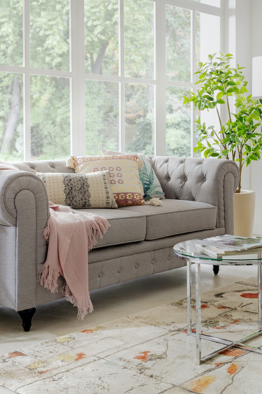 Sofá color gris con cojines estilo boho y una manta rosada. Mesa de centro redonda, planta en una maceta blanca y gran ventanal sin cortinas.