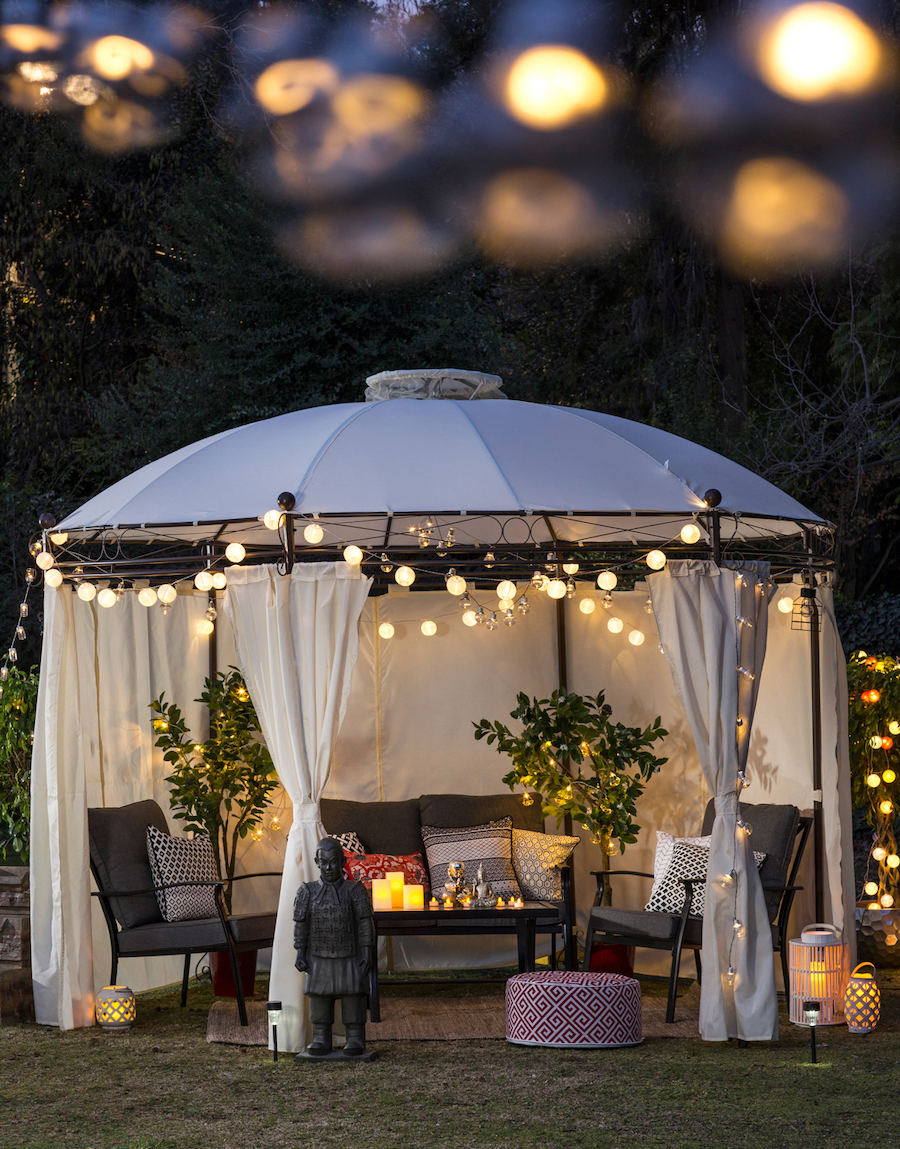 Terraza de invierno, con gran toldo blanco con techo curvo y cortinas abiertas. Dentro hay un juego de living de terraza con cojines gris oscuro. Guirnaldas de luces encendidas