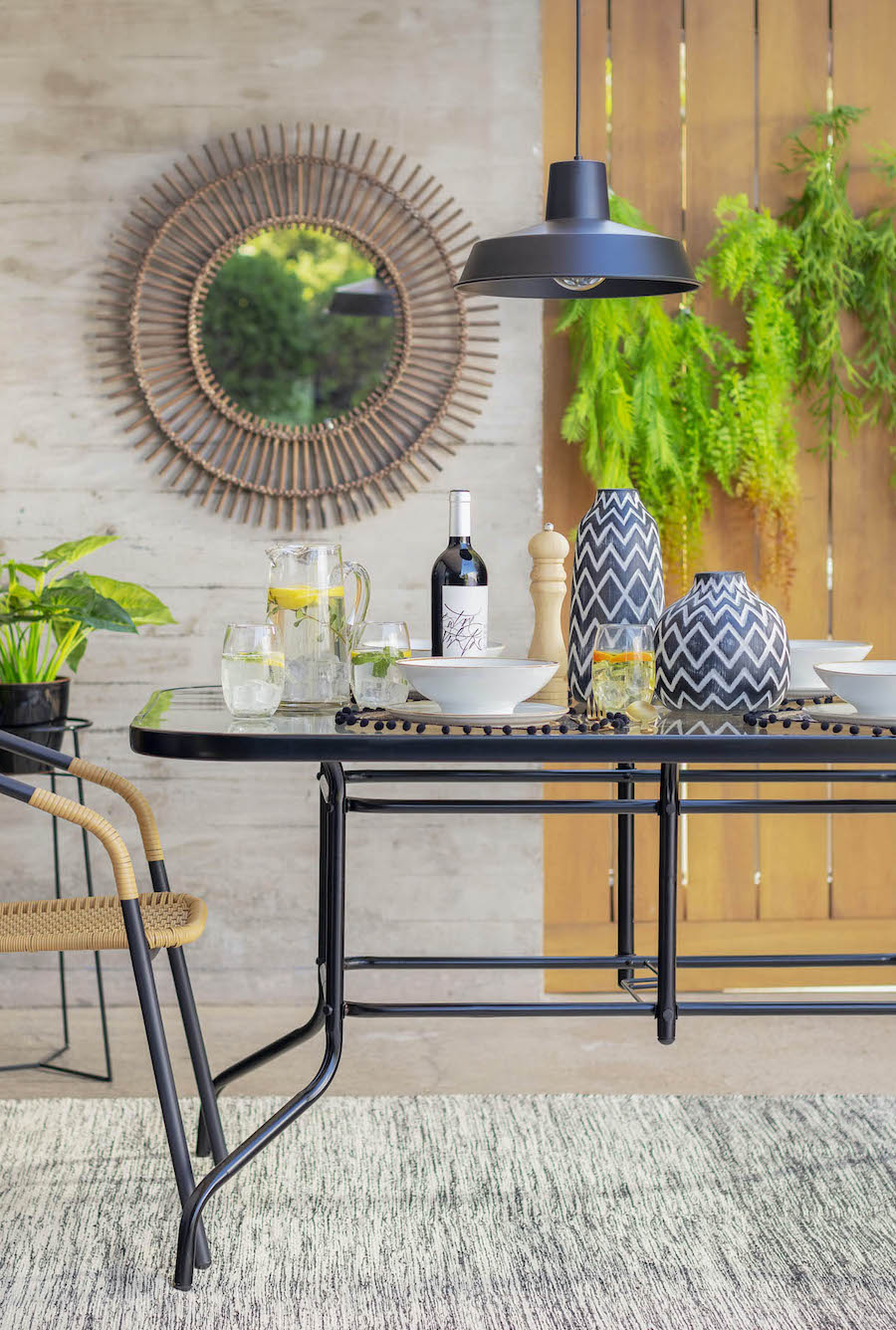 Comedor exterior con mesa de vidrio y sillas de metal y ratán. Sobre la mesa hay comida, botellas y jarrones de cerámica azul. Lámpara negra colgante y un gran espejo redondo en el muro