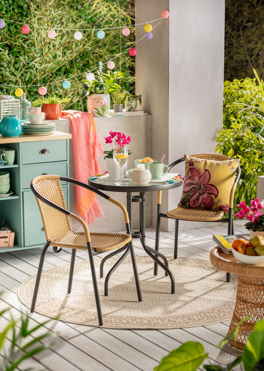 Terraza colorida con comedor de ratán y vidrio, para dos personas. Atrás hay un mueble tipo isla, color turquesa, con vajilla de colores. 