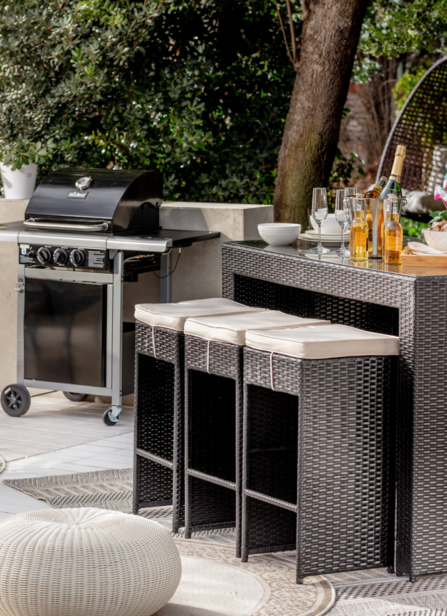 Terraza con comedor alto de ratán, color negro. Atrás se ve una parrilla de acero con tapa negra.