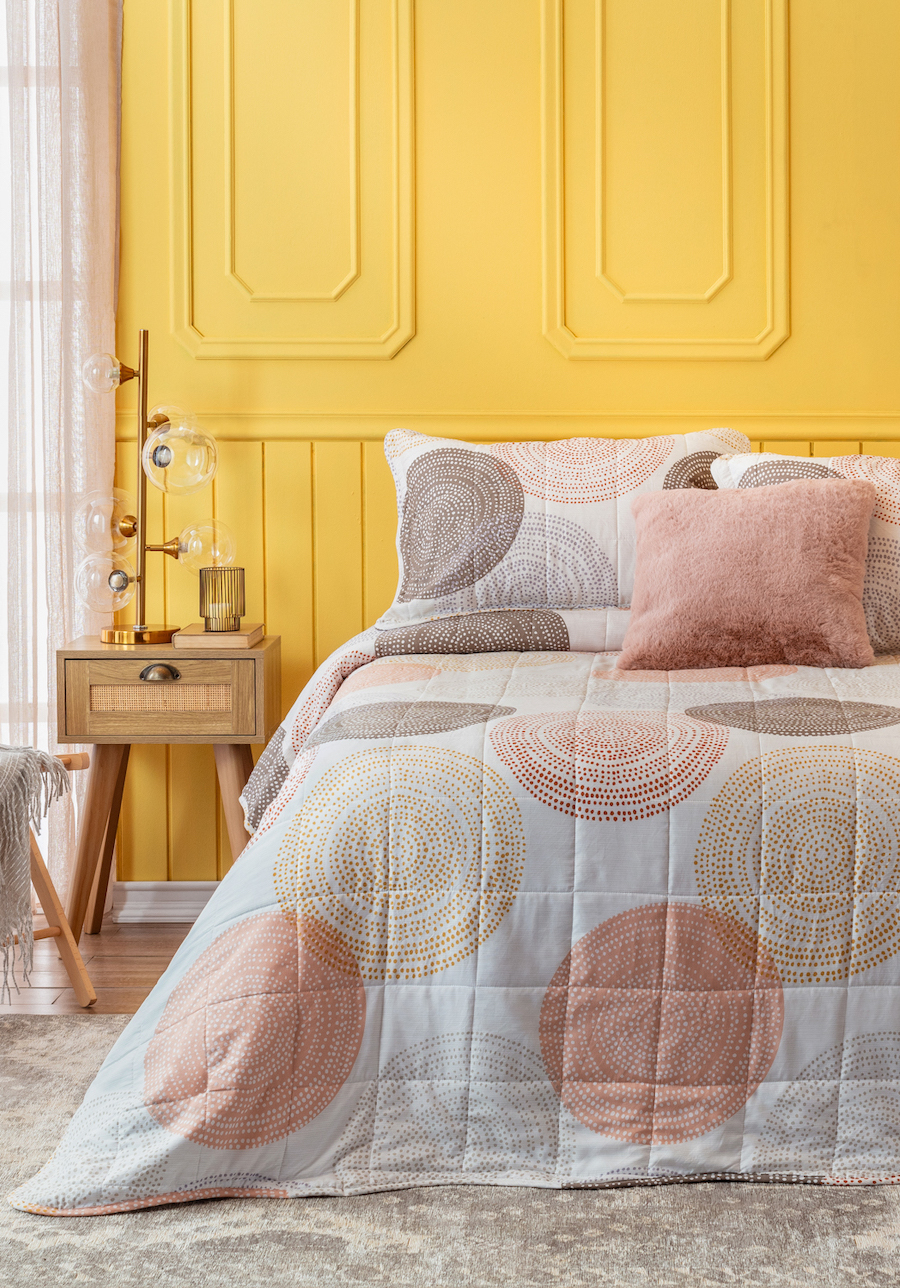 Dormitorio con gran contraste de colores. Muro amarillo con molduras y cama de dos plazas con plumón blanco con círculos café, rosa y beige. Velador de madera con un cajón