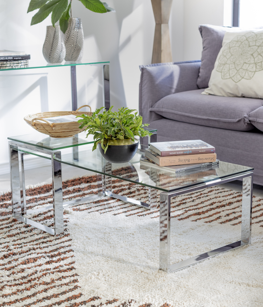 Mesa de estilo moderno, con patas vidriadas y base transparente de vidrio. Está sobre una alfombra con líneas cafés y azules. Sofá gris con cojín blanco.