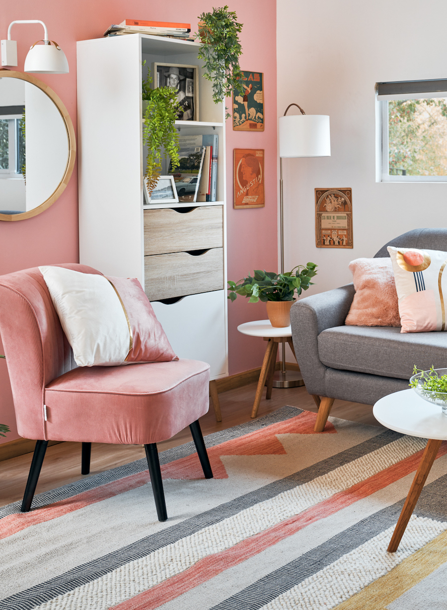 Living de estilo clásico con una pared de acento color rosa. Sofá gris y poltrona rosada. Repisa blanca pegada al muro junto a un espejo redondo