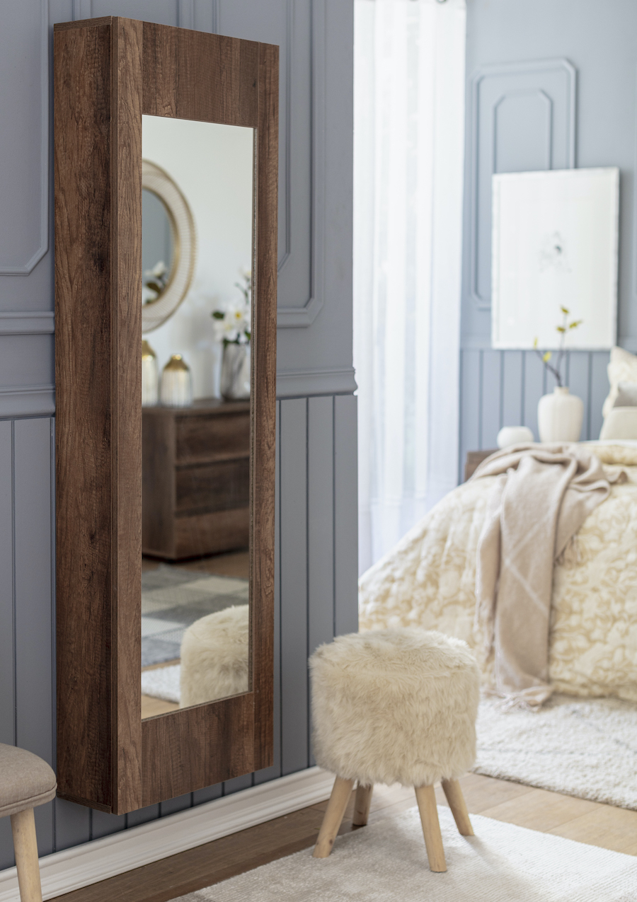 Espejo de cuerpo entero con marco de madera oscura colgado en la pared de una habitación con decoración elegante en tonos grises y neutros. Frente al espejo, hay un pequeño taburete redondo cubierto con tela de peluche.