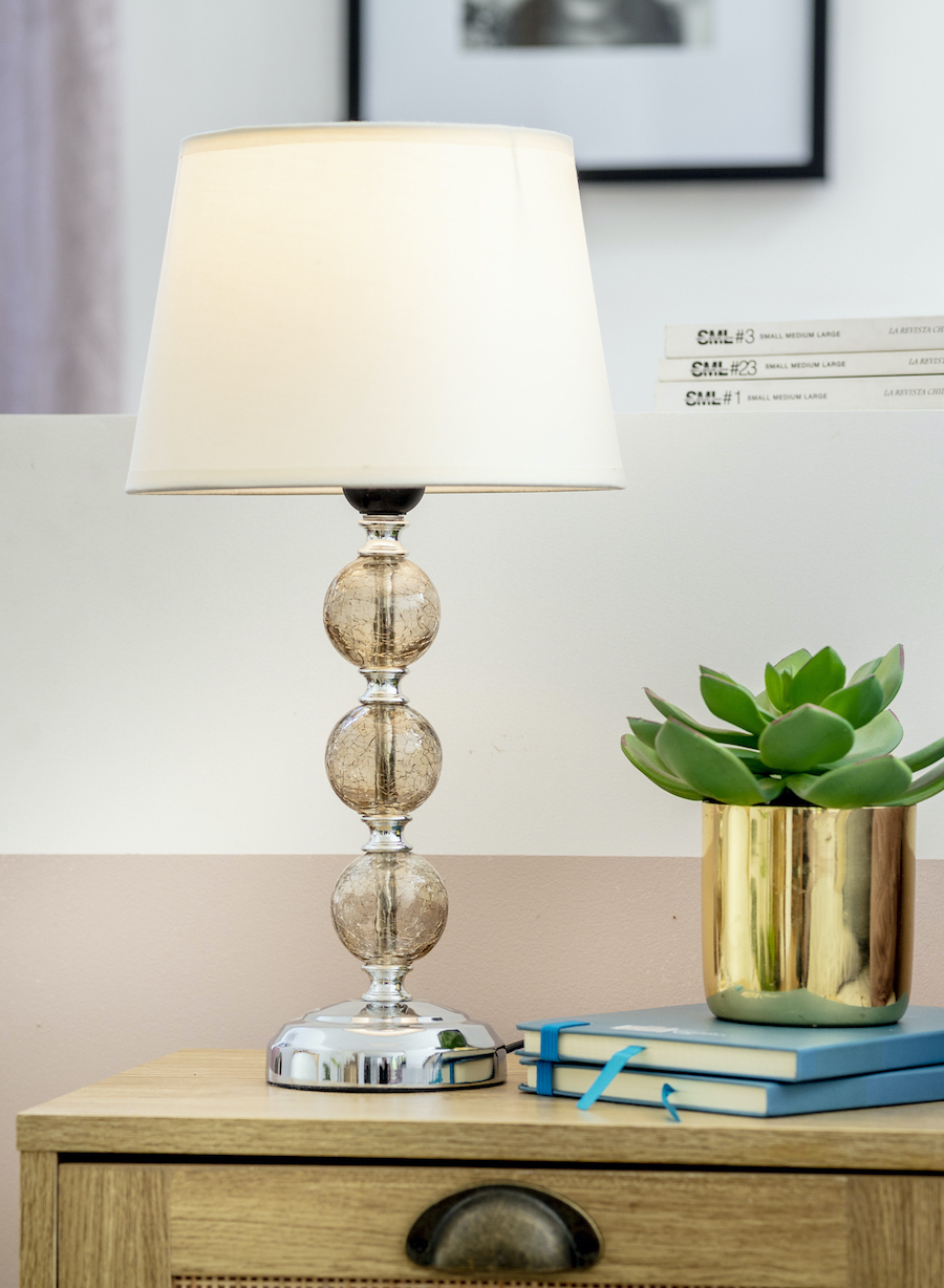 Lámpara de mesa decorativa con pantalla blanca y base esférica de cristal. Al lado hay un macetero dorado con una suculenta verde y dos libros apilados uno encima del otro. Al fondo hay libros.