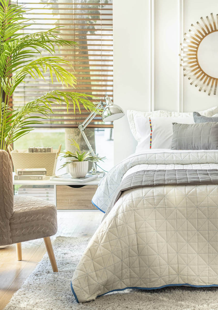 Dormitorio con una cama con una manta acolchada gris, almohadas blancas y una silla gris junto a una mesita de noche de madera con libros y una lámpara. Junto a la cama, junto a la ventana, hay una planta en una maceta.