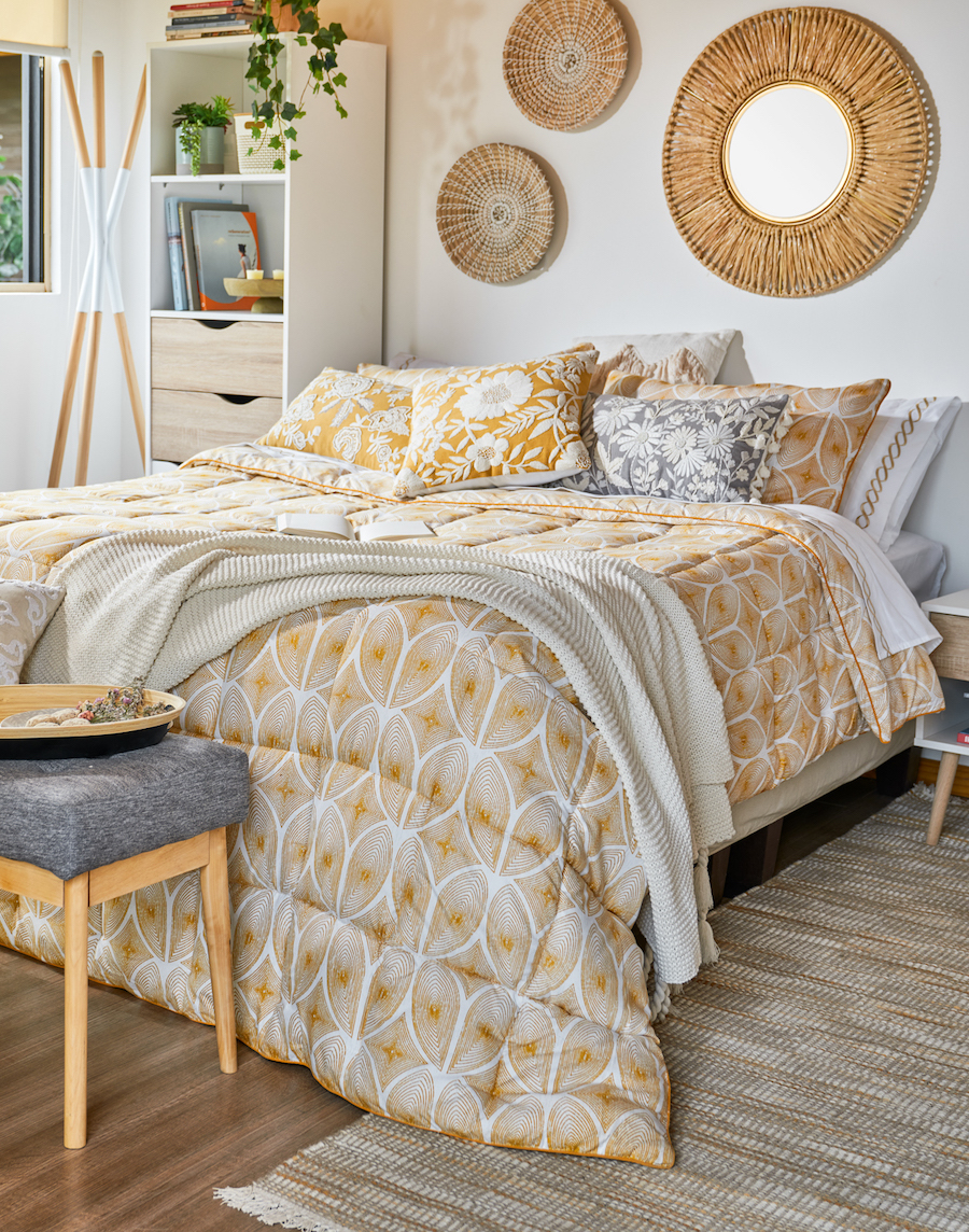 Dormitorio de estilo boho chic con una composición de espejos de marco de fibra natural sobre el respaldo de la cama. El cobertor tiene un diseño blanco con amarillo, al igual que algunos cojines.