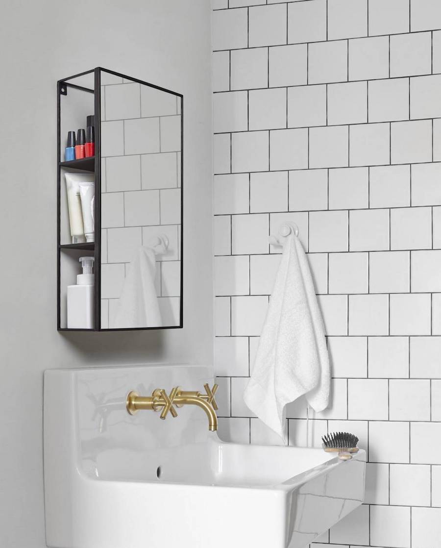 Baño de pared con cerámicas blancas rectangulares, un lavamanos con grifería dorada y un espejo de marco negro que detrás esconde un botiquín.