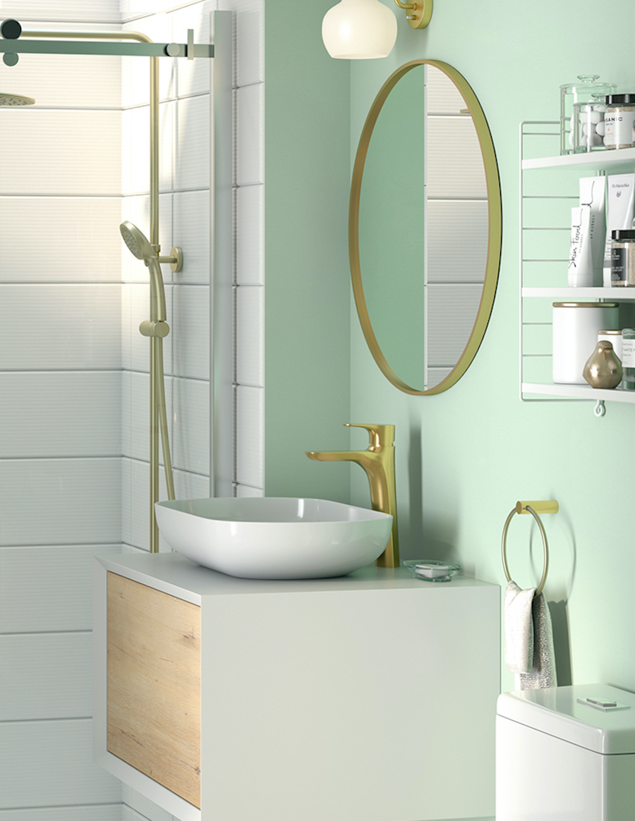 Baño de pared verde pastel y ducha con cerámica blanca. Hay un espejo redondo dorado sobre un mueble vanitorio blanco con puerta de madera, lavamanos blanco y grifería dorada. Al costado hay una repisa con distintos artículos de higiene y belleza.