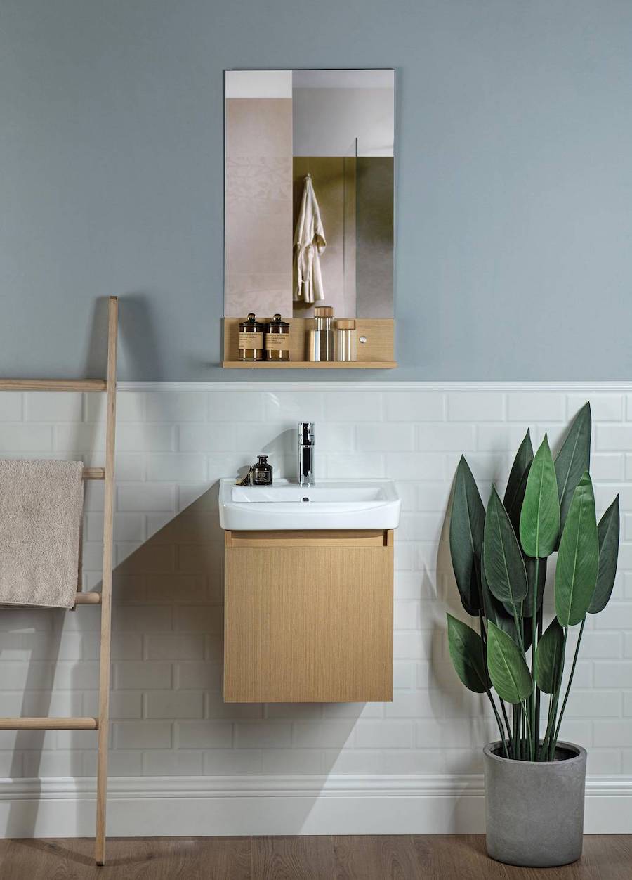 Baño de pared gris azulado y cerámicas blancas en la parte inferior. Hay un espejo con repisa de madera que hace juego con el mueble vanitorio que está debajo. Al costado hay una planta y una escalera decorativa con una toalla beige.