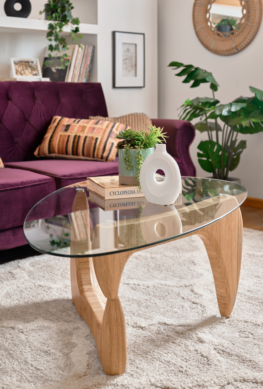 Detalle de una mesa de centro de vidrio ovalado con patas de madera. Sobre ella hay una pila de libros, un florero blanco y un macetero verde menta con una suculenta artificial. Al fondo hay un sofá morado con cojines en tonos tierra.