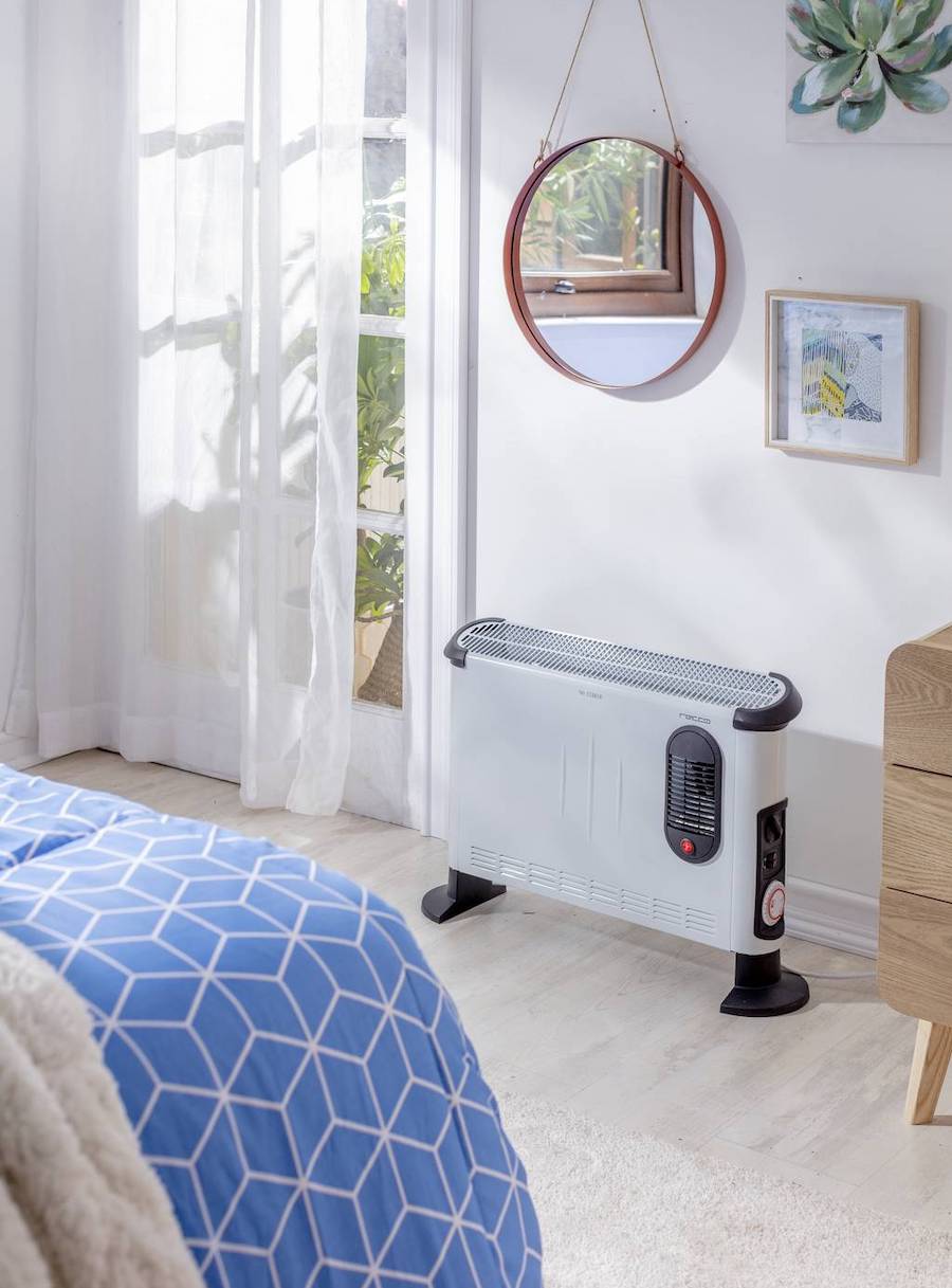 Convector eléctrico disponible en Sodimac, solo blanco con patas negras. Está en un dormitorio de estilo clásico frente a una cama con plumón azul.