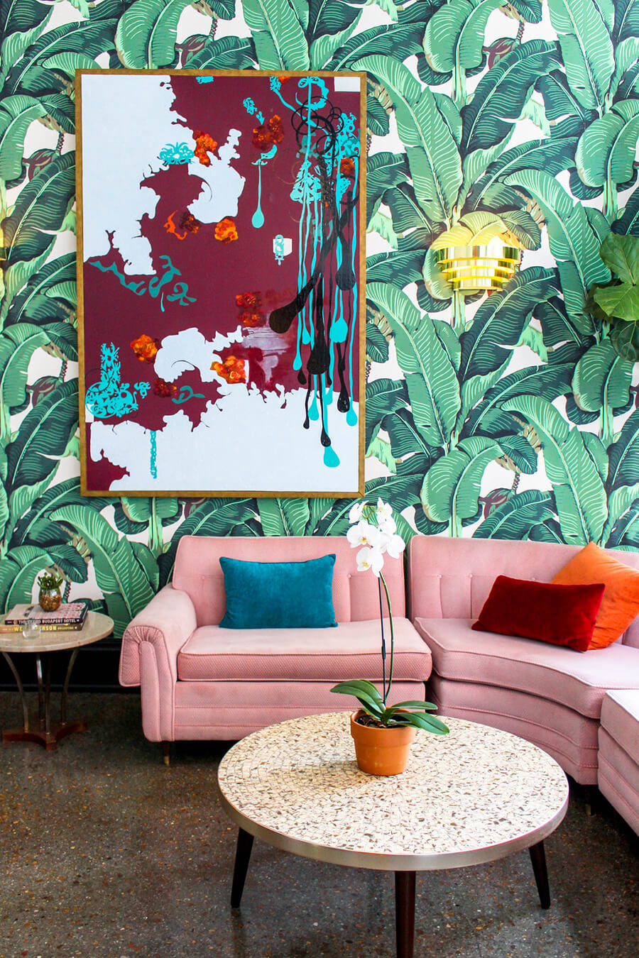 un living con sofá de varios cuerpos en color rosa, una mesa de centro circular y una pared con papel mural con detalles de plantas verdes y un cuadro