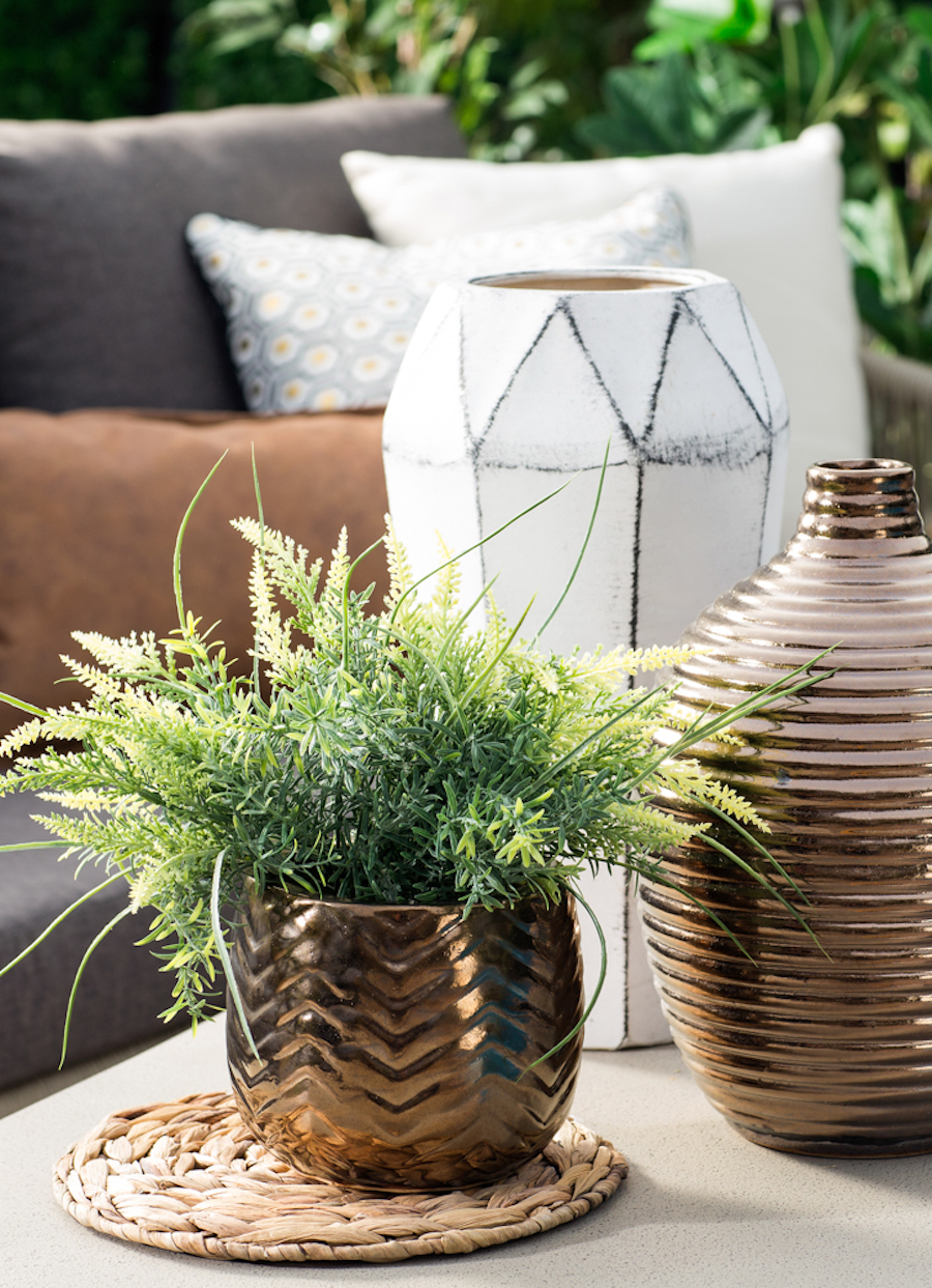 Detalle de una mesa de terraza con un macetero color cobre con una planta, junto a un jarrón de cobre y otro geométrico, de cerámica blanco.