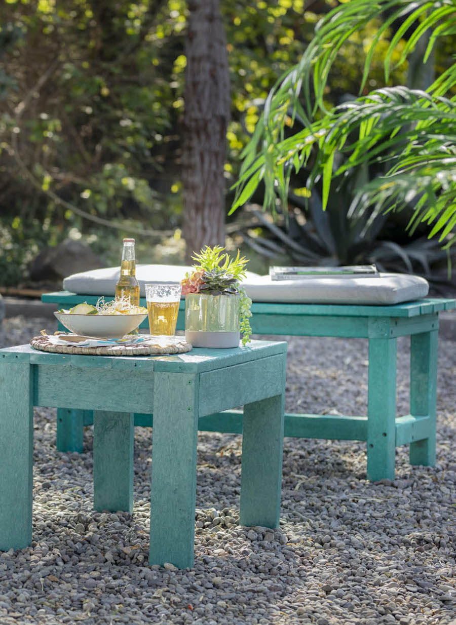 Terraza de madera color turquesa. Mesa y banqueta bajas sobre piedras, rodeadas de plantas y árboles