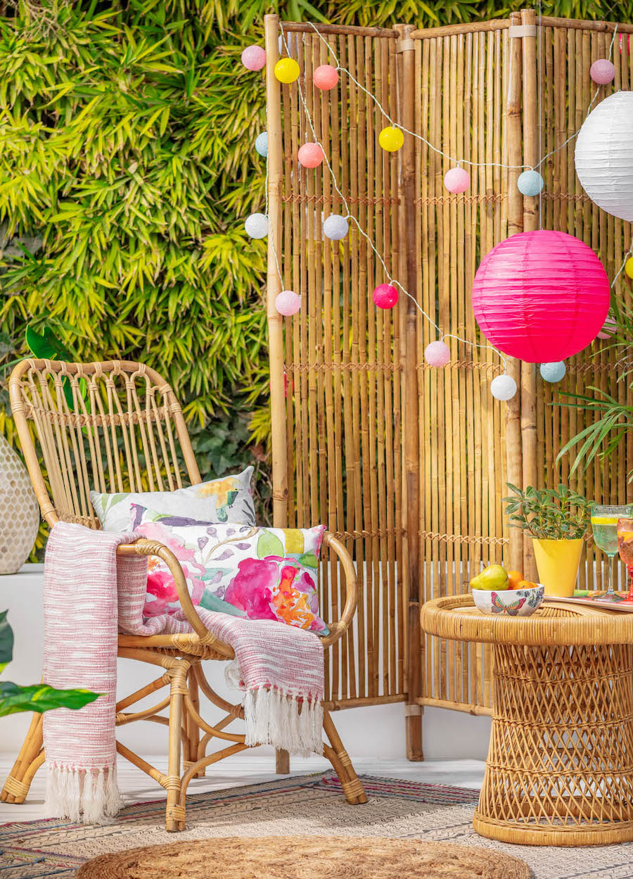 Pequeña terraza con biombo de bambú para privacidad. Silla y mesa de bambú con cojines de flores. Guirnaldas de luces redondas de colores vibrantes