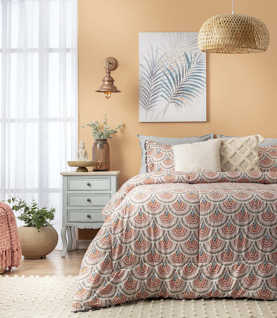 Dormitorio estilo boho chic. Cama de dos plazas con plumón bohemio, con patrón en tonos rosa y gris. Cojines beige. Velador estilo vintage con cajones. Gran cuadro sobre la cama con diseño de hojas.