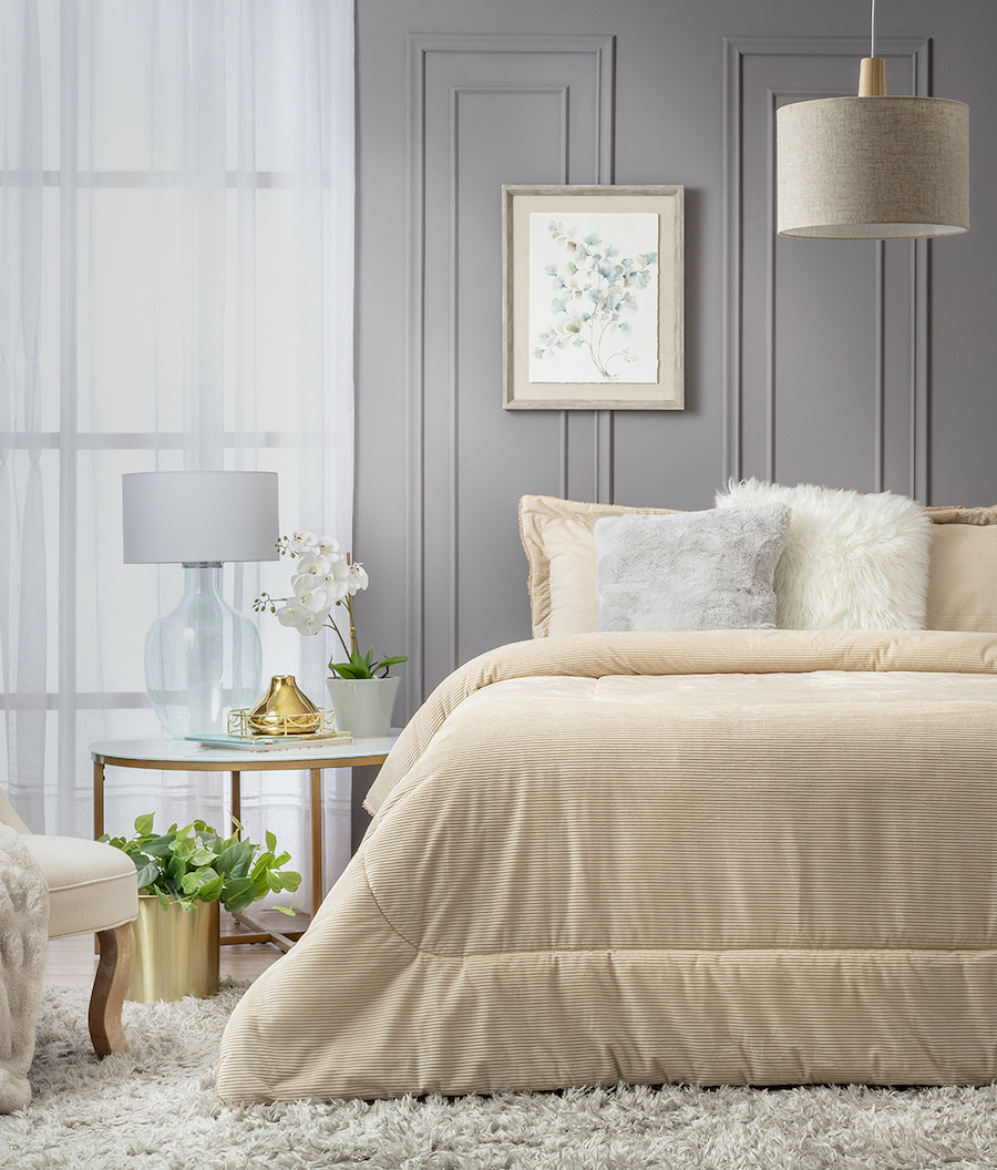 Dormitorio estilo clásico con cama de dos plazas. Plumón color beige y dos cojines peludos de color blanco y gris. Velador redondo con base de mármol, lámpara de mesa de vidrio y pantalla de tela.