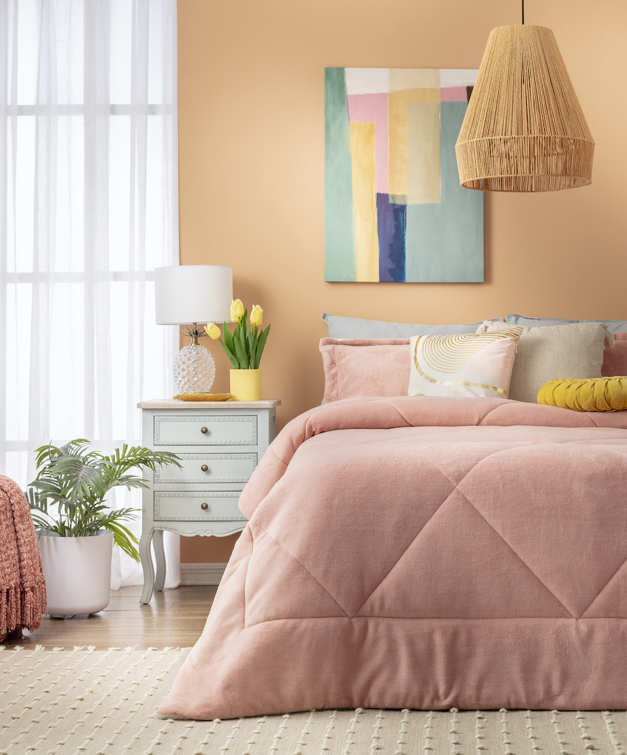 Dormitorio estilo dopamina. Cama de dos plazas con plumón color rosado. Cojines beige y amarillo. Gran cuadro abstracto como respaldo. Lámpara de techo de fibras naturales. 
