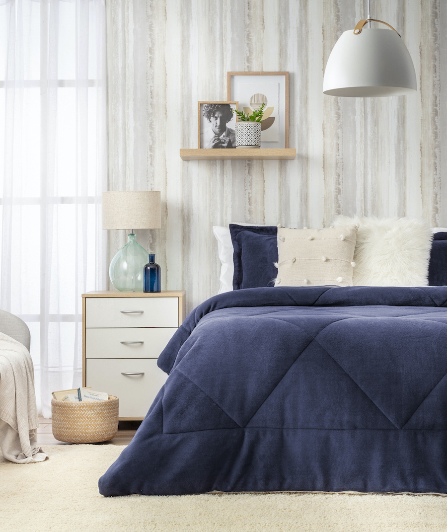 Dormitorio estilo scandifornian. Cama de dos plazas con plumón azul marino y cojines en color beige. Sobre la cama hay una repisa de madera flotante con cuadros y adornos. Velador moderno de color blanco con lámpara de vidrio y pantalla de tela