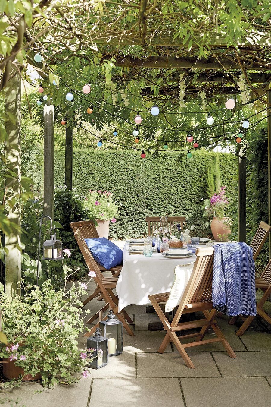 Terraza con treillage con plantas enredaderas y un comedor debajo. La mesa está cubierta con un mantel blanco y alrededor hay seis sillas de madera.
