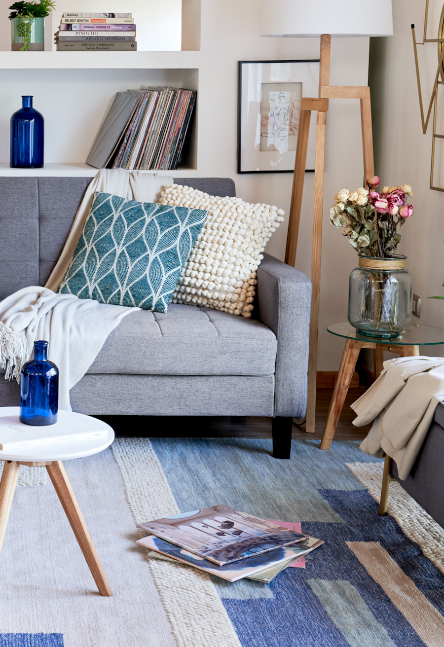 Sala de estar con un sofá gris con cojines blancos y calipso, una alfombra con distintos tonos azulados y sobre ella una pila de revistas.