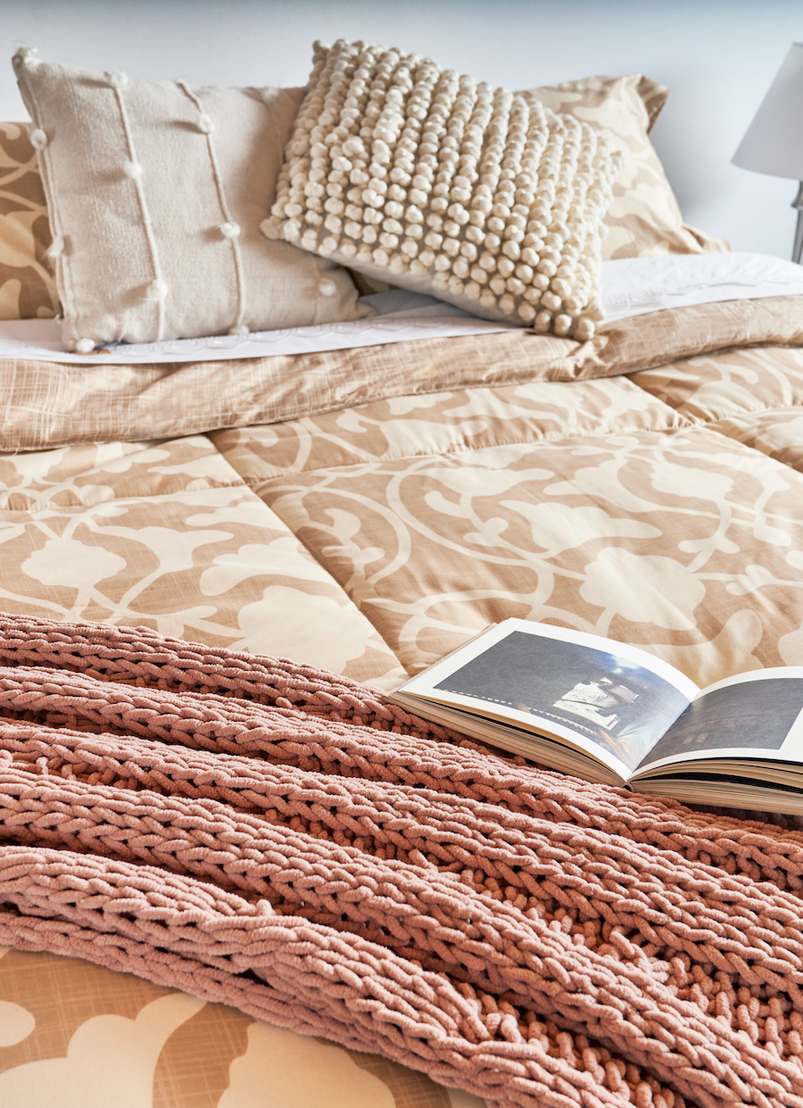 Detalle de una cama con un plumón con diseño en tonos crema, manta rosada y cojines beige.