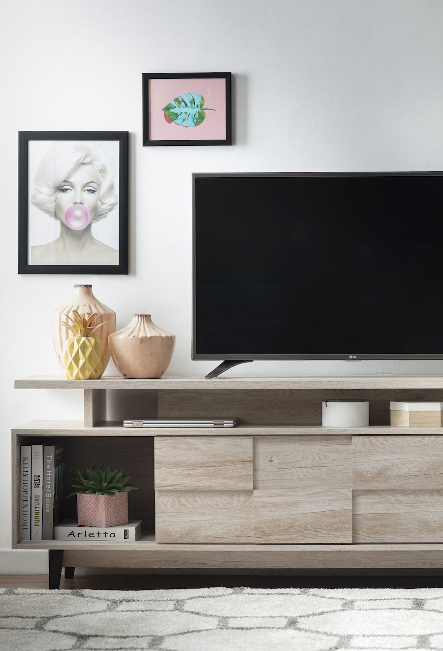 Rack de TV de madera clara con un televisor, jarrones en tonos amarillos y salmón. En el muro hay cuadros con ilustraciones y marco negro.
