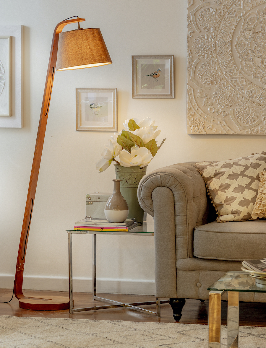 Detalle de un sofá gris en un living de paredes blancas y piso de madera. Junto al sofá hay una mesa lateral de vidrio con floreros sobre ella, y al costado, una lámpara de pie que emite luz cálida.