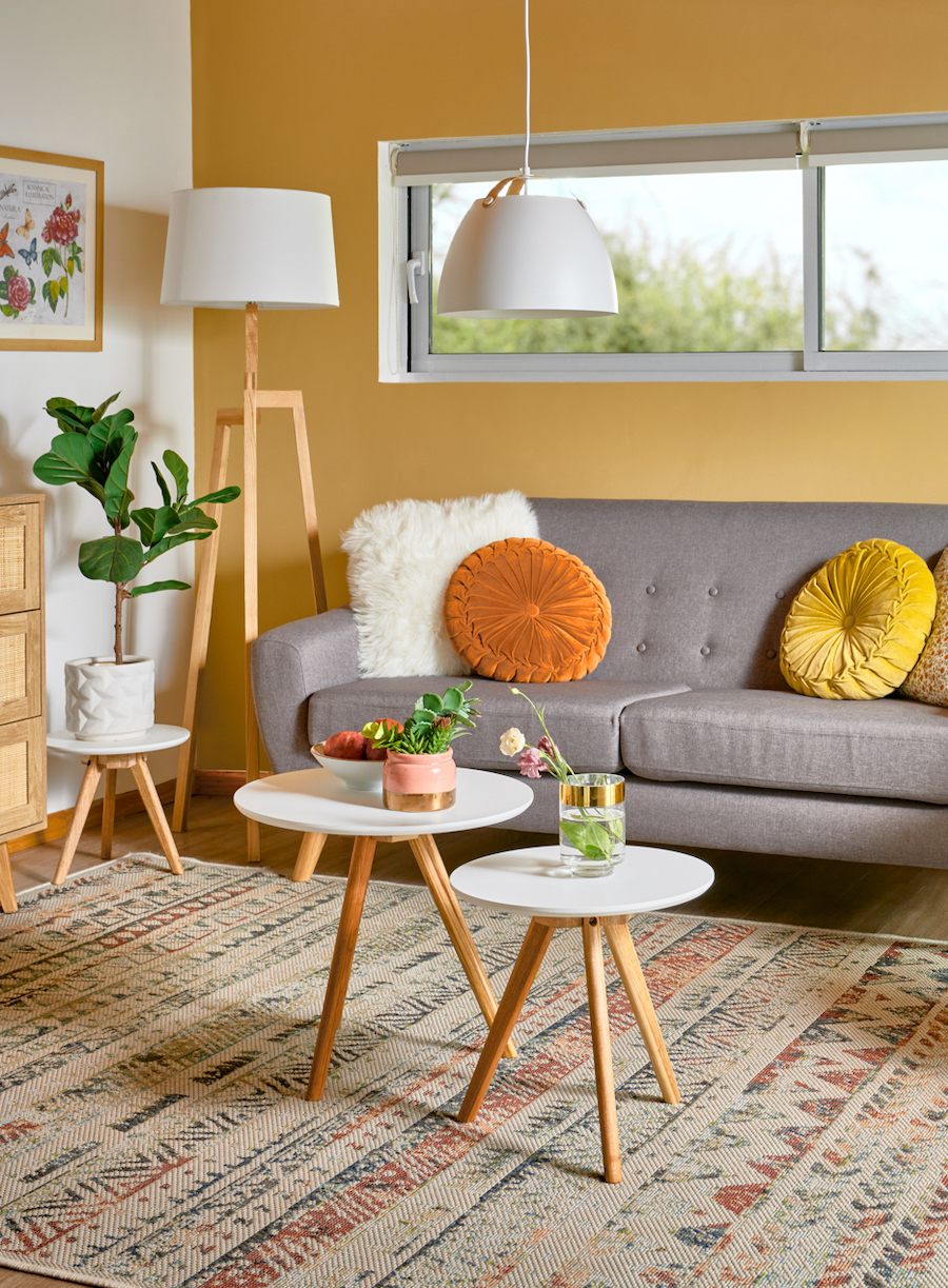 Living de paredes blancas y mostaza con una alfombra de colores tierra, un sofá gris con cojines en tonos amarillos y anaranjados. Frente a él hay 2 pequeñas mesas auxiliares que funcionan como mesa de centro. 