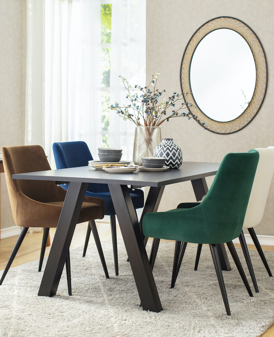 Comedor de muros beige, piso de madera y alfombra gris claro. La mesa de comedor es negra y alrededor hay 4 sillas con tapiz velvet de distintos colores, pero el mismo modelo. 