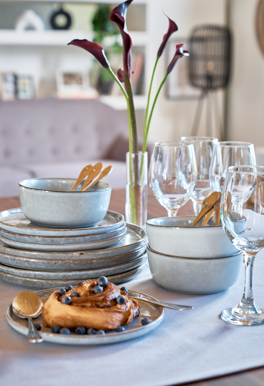 Detalle de una mesa de madera con vajilla color celeste. Platos y bowls apilados, copas y un plato con un rollo dulce. Pequeño florero de vidrio con flores.