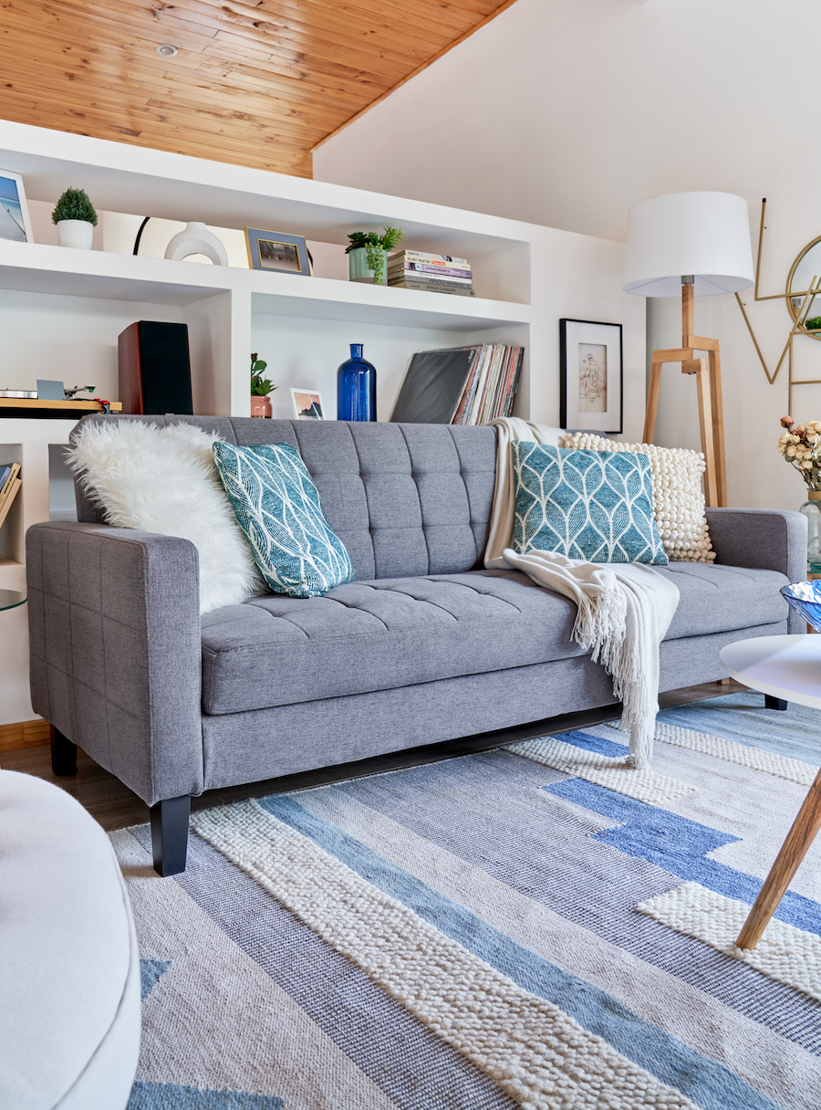 Living estilo clásico con sofá gris con cojines celestes y blancos. Alfombra en tonos azules, celestes y grises. Atrás una gran repisa empotrada con adornos y libros.