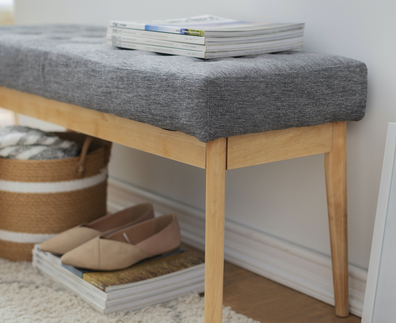 Detalle de una banqueta de tela gris con patas de madera. Abajo hay un canasto, revistas y unos zapatos.