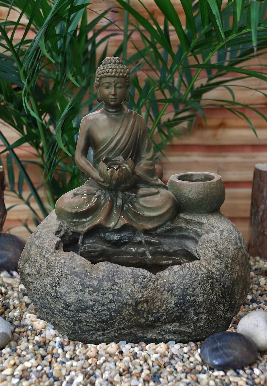 Rincón zen con pileta de agua que incluye la imagen de un buda. La fuente es de piedra, de olor gris oscuro. Está sobre pequeñas piedras y atrás se ven plantas.