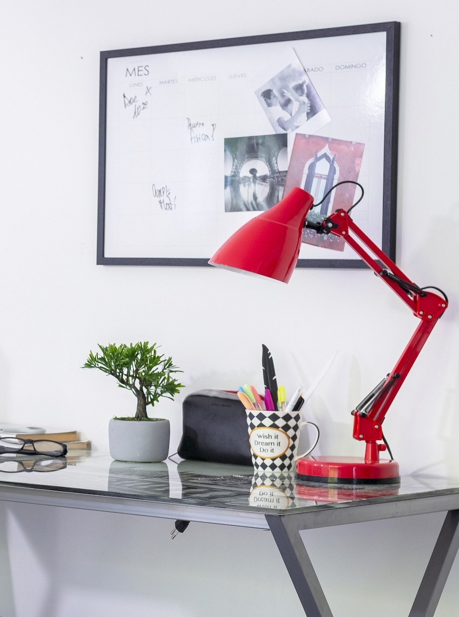 Mesa de escritorio con la teoría del rojo inesperado. Mesa metálica con base de vidrio, sobre ella una lámpara metálica roja, un vaso con lápices y una pequeña planta. En el muro una pizarra con marco negro