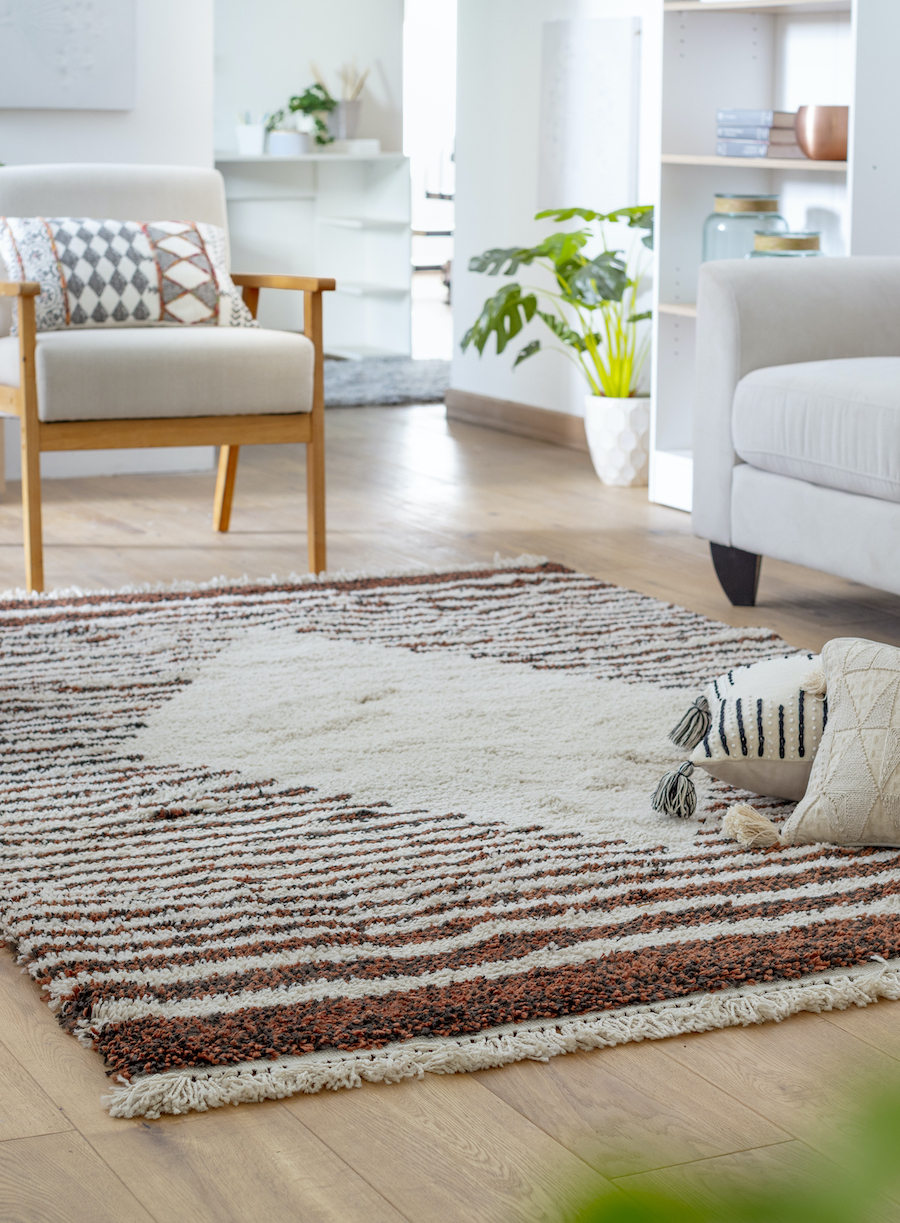Detalle de una alfombra de rayas blancas con café con un gran rombo blanco en el centro. Sobre ella hay dos cojines blancos y al costado, un sofá blanco invierno y una poltrona de madera.