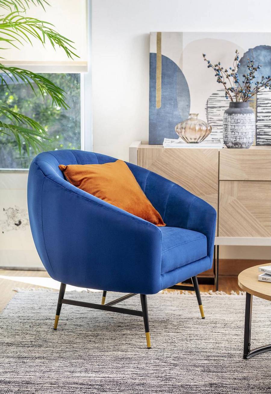 Poltrona azul con un cojín anaranjado en una sala de estar en tonos neutros, sobre una alfombra de color crema y delante de un buffet de madera clara.
