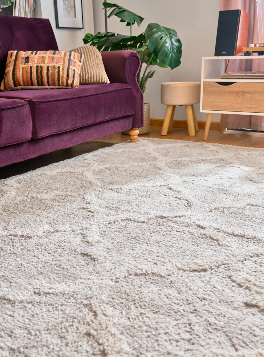 Detalle de un living con un sofá morado con cojines en tonos crema y una gran alfombra en color crema con un diseño de líneas blancas.