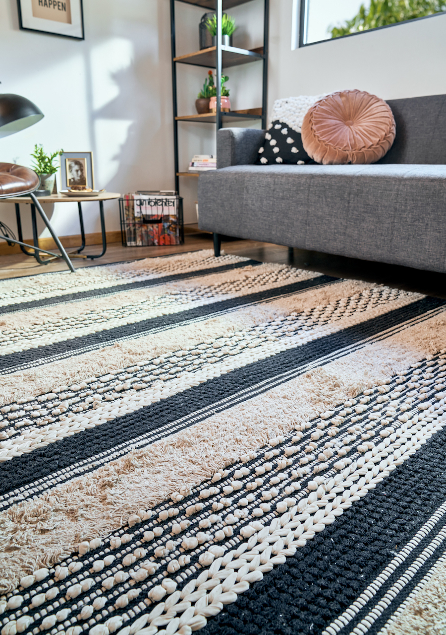 Detalle de una alfombra de lana. Es un tipo de alfombra con líneas blancas, negras y color crema, estilo ético. También hay un sofá gris y un sillón de estilo industrial de cuero café con patas metálicas negras.