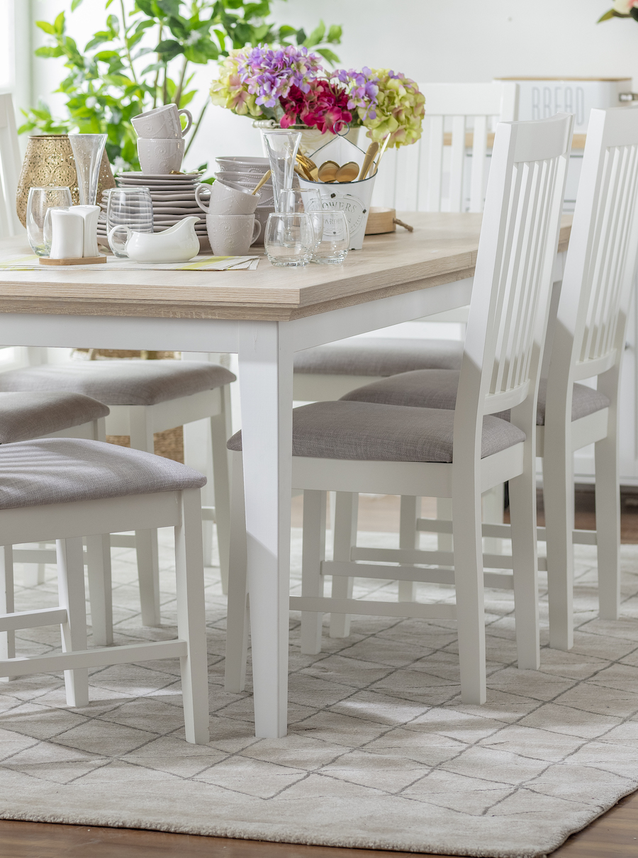 Comedor con mesa y sillas de madera blanca sobre un tipo de alfombra shaggy de color blanco con líneas grises. Sobre la mesa hay menaje, desde platos hasta tazas en tonos neutros.